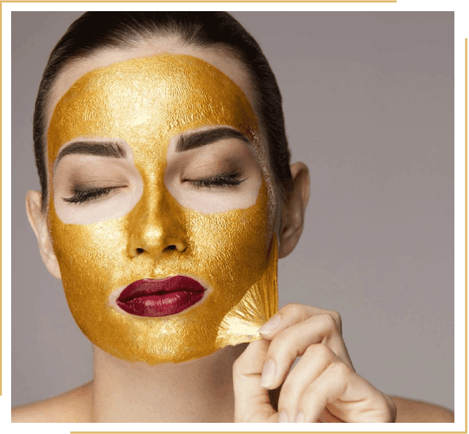 Woman applying a gold-colored facial mask, with her eyes closed and wearing dark red lipstick, in a close-up shot.