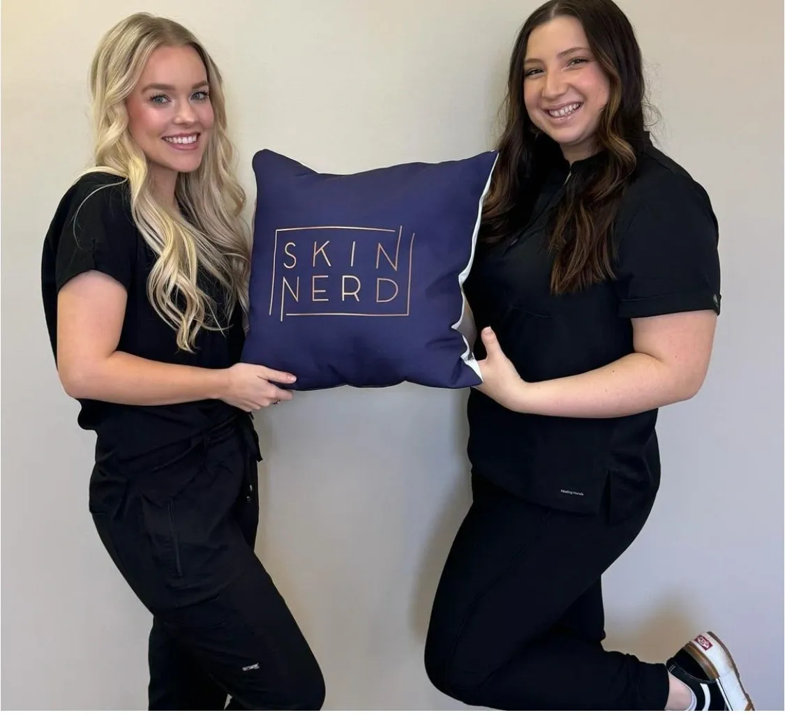 Two women in black outfits holding a navy pillow with "Skin Nerd" printed on it, standing against a plain wall.